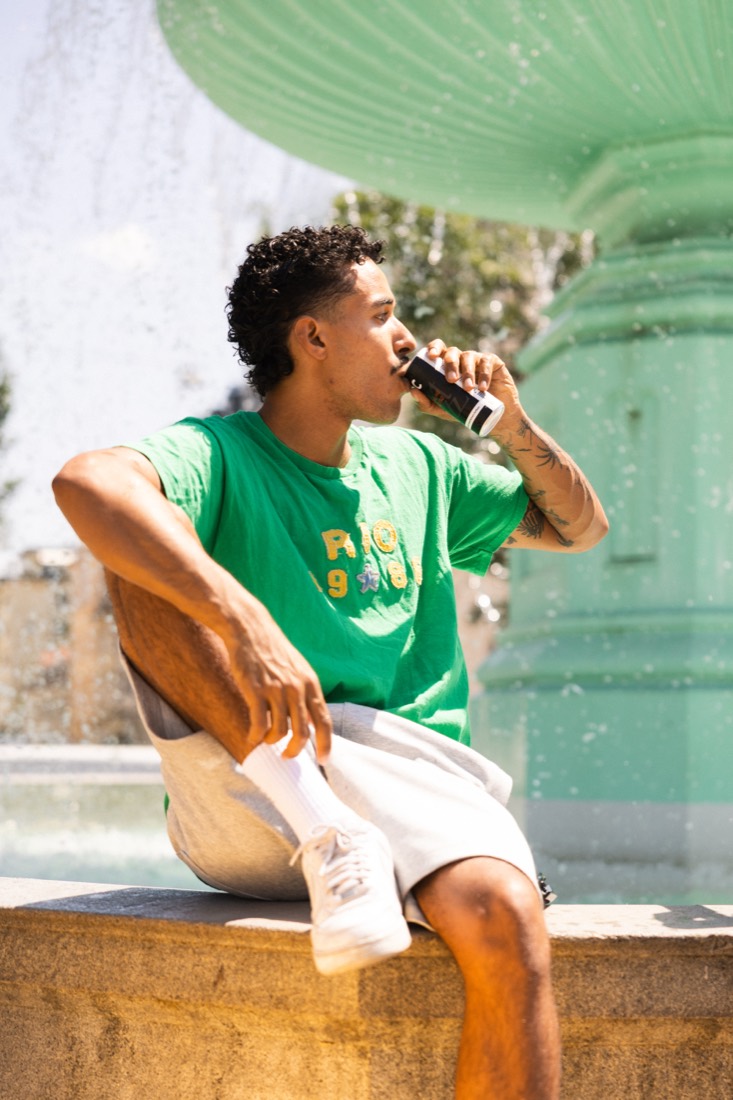Mann im grünen Shirt trinkt Espresso Tonic am sonnigen Brunnen – Foto: forStory